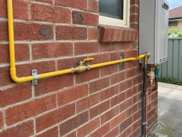 Yellow gas pipe and fittings mounted on a brick exterior wall beside a small gas unit in Lidcombe